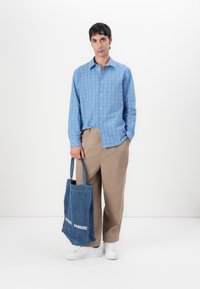 Blau karierter Hemd mit Kragen, beige Hose und weiße Sneaker. Hält eine Denim-Tote-Tasche mit der Aufschrift "SAMØE SAMØE".