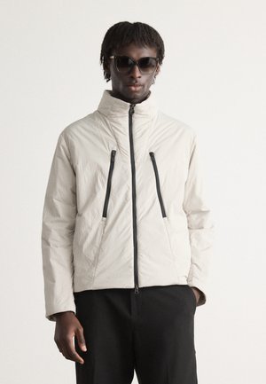 Light gray puffer jacket with a high collar, two black zippered pockets, and a textured finish, worn over dark trousers. Black sunglasses included.