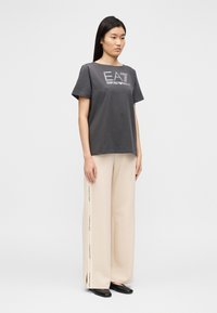 Szara bawełniana koszulka z krótkimi rękawami i okrągłym dekoltem, z białym logo "EA7 Emporio Armani" zdobionym lśniącymi akcentami. Beżowe spodnie o szerokich nogawkach.