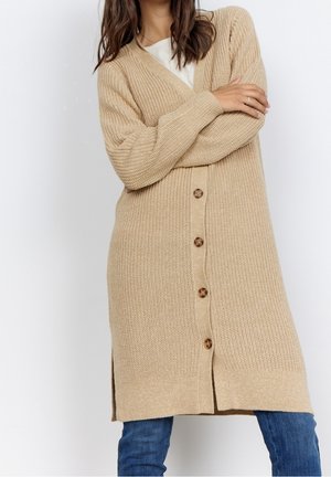 Femme portant un long cardigan beige en maille avec quatre boutons, un haut blanc et un jean bleu, bras croisés devant un fond uni.
