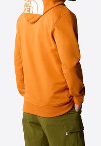 Persona che indossa un felpa arancione con un grande logo bianco sul cappuccio e pantaloni cargo verde oliva, con le mani in tasca, di spalle.