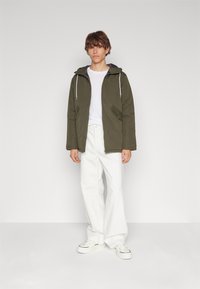 Veste à capuche vert olive avec doublure grise, deux poches latérales, cordon de serrage, coupe décontractée, associée à un pantalon large blanc et des baskets blanches.