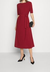 Rotes Midi-Kleid mit kurzen Ärmeln und Taillengürtel, aus strukturiertem Stoff. Kombiniert mit schwarzen spitzen Absätzen mit goldenen Akzenten.
