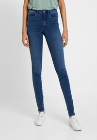 Calças skinny de jeans azul de cintura alta com uma textura suave, dois bolsos frontais e fecho de botão, combinadas com ténis brancos.