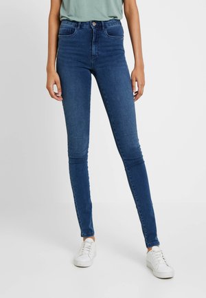 Calças skinny de jeans azul de cintura alta com uma textura suave, dois bolsos frontais e fecho de botão, combinadas com ténis brancos.