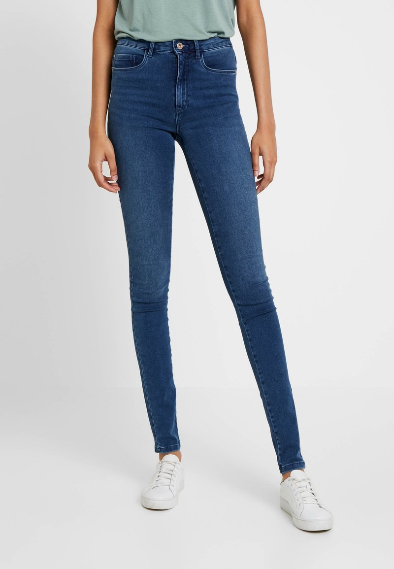 Calças skinny de jeans azul de cintura alta com uma textura suave, dois bolsos frontais e fecho de botão, combinadas com ténis brancos.