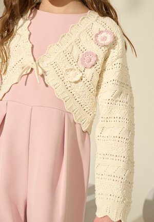 Dziecko ubrane w jasnoróżową sukienkę z kremowym bolerkiem na szydełku, ozdobionym falistymi brzegami i aplikacjami kwiatów w różowym i białym kolorze.