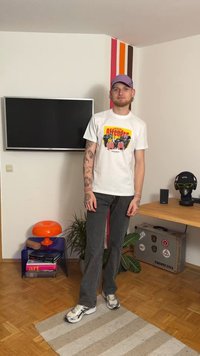 Valkoinen graafinen t-paita, jossa värikäs printti, tummat farkut ja harmaat lenkkarit. Seisoo huoneessa, jossa on televisio ja koriste-esineitä.