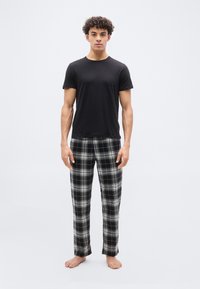 Νεαρός άνδρας που στέκεται ξυπόλητος φορώντας μαύρο μπλουζάκι και ασπρόμαυρο καρό παντελόνι πιτζάμας σε λευκό φόντο.