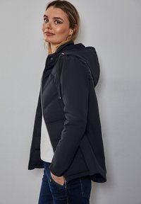 Svart pufferjacka med huva, har dragkedja framtill, elastiska �ärmmuddar och sidofickor. Tyget ser texturerat och varmt ut.