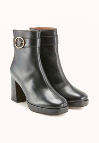 Bottes de cheville en cuir noir avec un talon bloc, présentant un accent circulaire métallique et une finition lisse, conçues pour une silhouette ajustée.