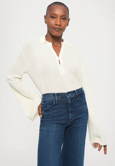 Donna con i capelli corti che indossa un maglione a polo bianco a maniche lunghe e jeans blu a vita alta, in posa con una mano nella tasca.