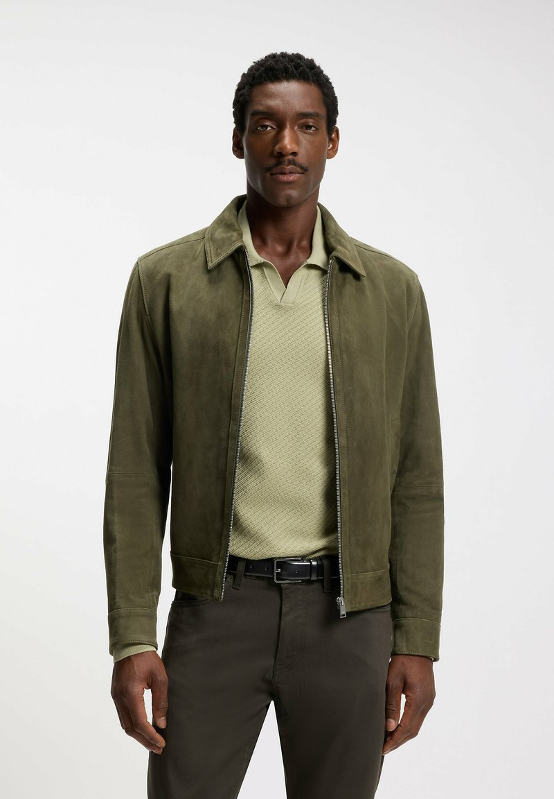 Homme portant une veste en daim vert olive sur un polo vert clair texturé et un pantalon foncé, se tenant devant un fond blanc uni.