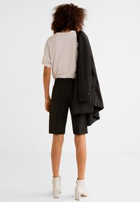 Shorts noirs à rayures avec un t-shirt gris clair. Un blazer noir est drapé sur un bras, accompagné de bottes beiges montantes.