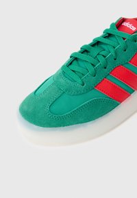 Rohelise seemisnahast ja sünteetilise materjaliga toss, millel on punased aktsendid, ümmargune varvas ja läbipaistev kummist tald. Tossu paelad on sama rohelise värvusega.