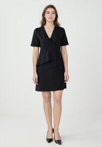 Robe noire avec un design enroulé, manches courtes et un détail de volants en couches asymétriques à la taille. Tissu lisse avec une coupe structurée.