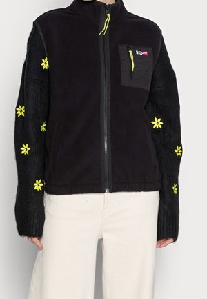 Sort lynlåstrøje med strikkede ærmer, dekoreret med gule broderede blomster, kombineret med off-white bukser.