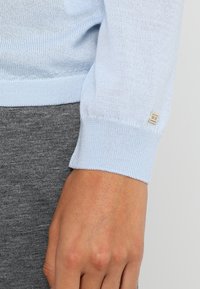 Jasnoniebieski sweter z dzianiny, z rękawem zakończonym ściągaczem, z małym haftowanym logo. Widać szary materiał poniżej. Gładka struktura.