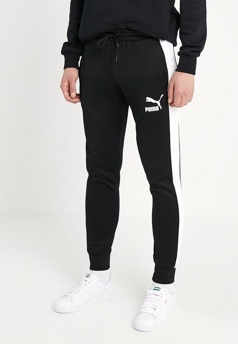 Homme portant un pantalon de survêtement noir Puma avec des bandes blanches latérales et des baskets blanches Puma, debout devant un fond uni.