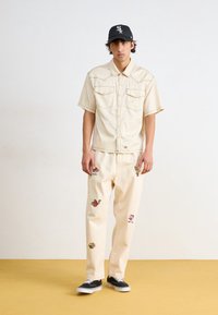 Beige short-sleeve shirt with two front pockets, paired with cream pants featuring colorful embroidered designs. Black cap and black sneakers.