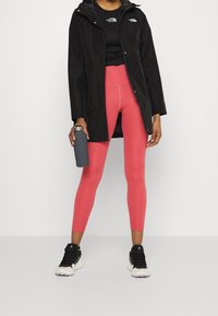 Black waterproof jacket over a black fitted top, paired with bright pink leggings and black athletic shoes. A gray water bottle is held in one hand.