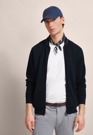 Navy zip-up jacket with textured fabric, worn over a white t-shirt. Gray trousers and a navy cap, with a striped scarf around the neck.