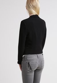 Černý přizpůsobený blazer s krátkým střihem, se hladkým povrchem a ostrými klopami. Nošený s světlými šedými džíny se zipovými detaily.