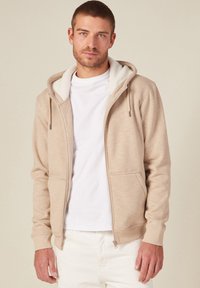 Felpa beige con zip, foderata in morbido pile bianco, con tasche a marsupio e cappuccio regolabile. Indossata sopra una t-shirt bianca, presentata su uno sfondo neutro.