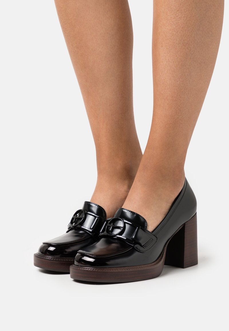 Mocassins à talons carrés en cuir noir brillant avec détail de boucle ronde portés aux pieds sur fond blanc.
