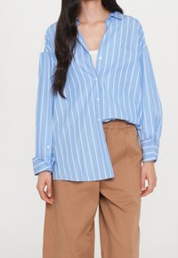 Niebieska i biała w paski koszula z guzikami, z kieszenią na piersi, lekko oversize'owa, z podwiniętymi rękawami, w zestawie z beżowymi spodniami.