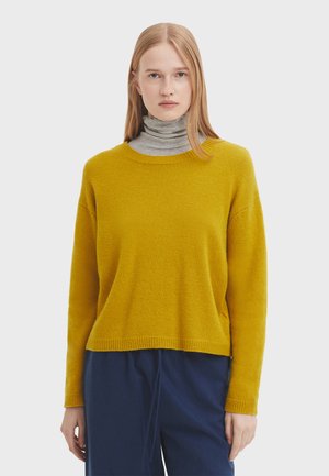 Pull en maille jaune moutarde avec un col rond, des manches longues et une coupe décontractée, associé à un col roulé gris en dessous.