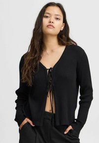 Svart ribbad cardigan med V-hals och snörning, med långa ärmar och avslappnad passform. Mjuk textur och minimalistisk design.