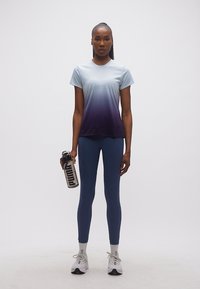 Γυναίκα με μπλε αθλητικό μπλουζάκι σε αποχρώσεις, σκουρόχρωμο μπλε κολάν, λευκά αθλητικά παπούτσια, κρατώντας ένα διαφανές παγούρι Puma, στέκεται μπροστά σε απλό φόντο.