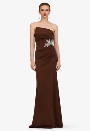 Woman wearing a strapless dark brown gown with silver floral embellishment on the waist, standing against a plain light background.