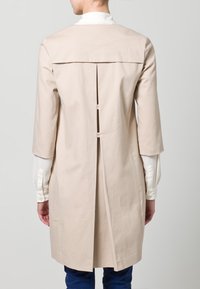 Beige puuvillatakki, jossa on kolmasosa hihat, takasaumassa laskostus ja napit. Ominaispiirteitä ovat puhdas ja minimalistinen muotoilu.