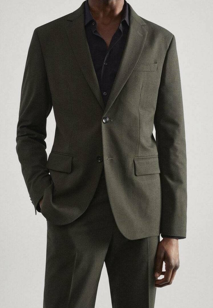 Homme en costume vert olive foncé avec chemise noire, une main dans la poche, debout devant un fond clair uni.