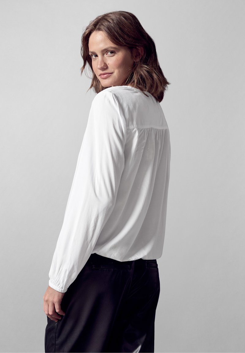 Weißes Langarmshirt mit lockerer Passform und gerafftem Rückendetail, getragen mit schwarzen Hosen; glatte Textur, einfaches Design, keine sichtbaren Muster.