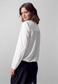 Witte blouse met lange mouwen, een geplooide beleg en elastische manchetten, gecombineerd met zwarte broek, weergegeven tegen een neutrale achtergrond.