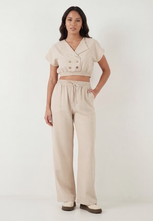 Žena u bež krojenom dvorednom topu s elastičnim strukom i odgovarajućim širokim hlačama s vezicom, stoji s rukom u džepu.