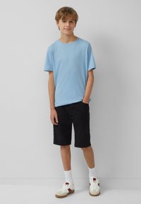 Lichtblauwe katoenen T-shirt gecombineerd met zwarte shorts, witte sokken en sneakers met beige en rode accenten. Eenvoudige, casual outfit.