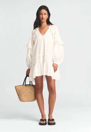 Hvid, knælang kjole med flæser på ærmerne og kanten. Båret med sorte sandaler og holder en vævet stråtaske. Glat tekstur, afslappet design.