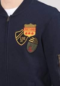 Sudadera azul marino con parches bordados en amarillo, rojo y verde. Los parches incluyen 'FIESTA DURO', 'VIVE LA VIDA' y 'SÉ LIBRE'. Detalle de cremallera incluido.