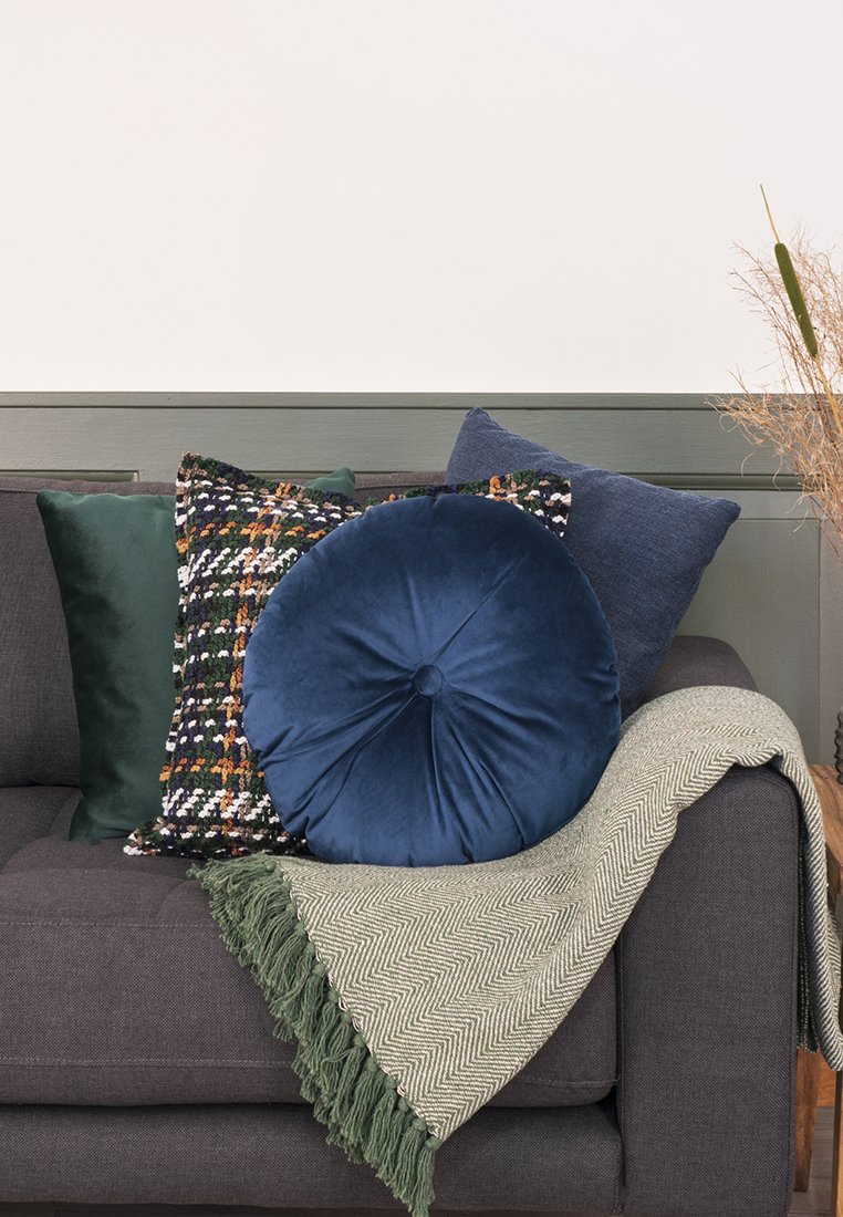 Graues Sofa mit grünen, multicolor, blauen runden und blauen quadratischen Kissen sowie einer grünen, fransigen Decke auf der Armlehne.