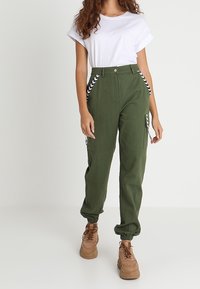 Olivgrön cargobyxor med sidofickor, elastiska ankelslut och kontrasterande svartvita zigzagdetaljer. Kombineras med en vit t-shirt.