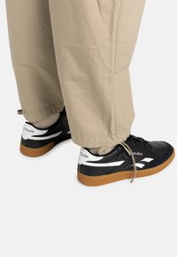 Sort og hvide Reebok sneakers med en tekstureret læderoverdel, gummisål og gummisål. Båret med lysebrune baggy bukser.