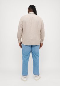 Camicia beige a maniche lunghe con colletto, dettaglio sulla spallina posteriore e polsini con bottoni, abbinata a jeans in denim blu chiaro e scarpe da ginnastica bianche.