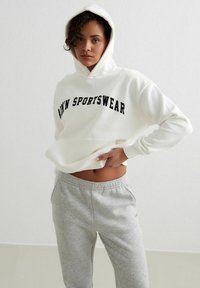 Sudadera blanca con "DWN SPORTSWEAR" en letras negras, con un bolsillo frontal y capucha, combinada con pantalones de chándal gris claro.
