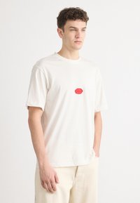 Hvid bomuldstrøje med et rundt rødt logo p�å brystet, med korte ærmer og en afslappet pasform. Stoffet ser blødt og glat ud.