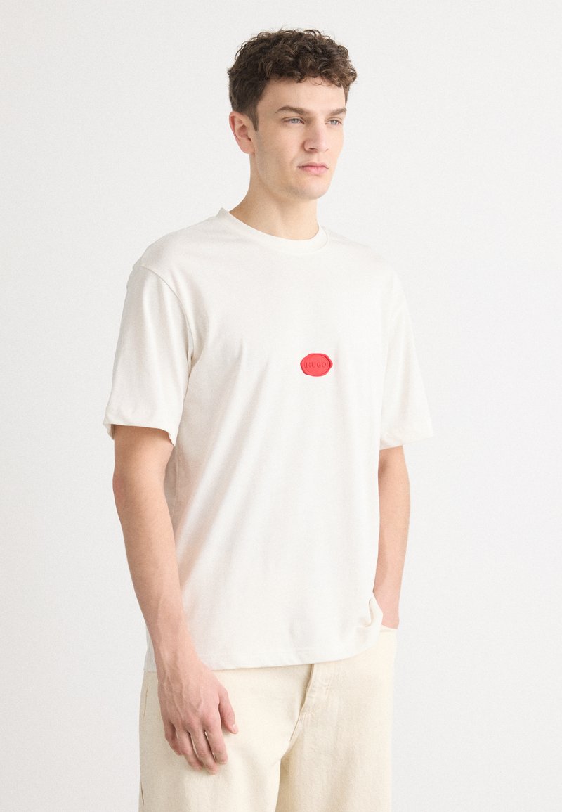 Vit bomull t-shirt med en rund röd logotyp på bröstet, med korta ärmar och en avslappnad passform. Tyget verkar mjukt och slätt.