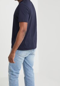 Granatowa koszulka z krótkim rękawem o gładkiej fakturze, w połączeniu z jasnoniebieskimi dżinsami. Koszulka ma prosty, okrągły dekolt.